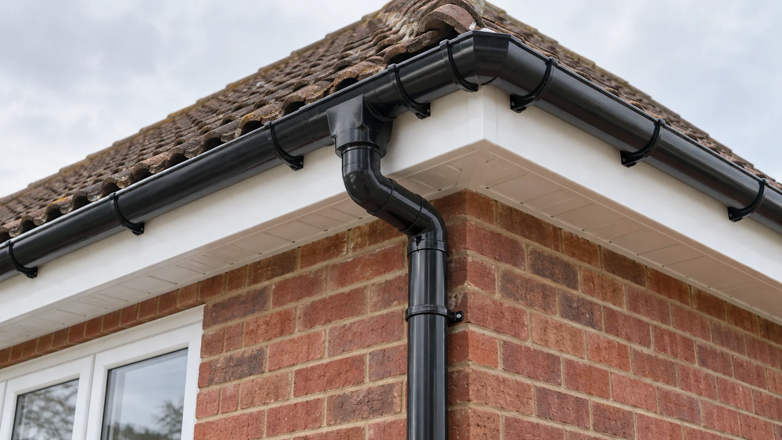 Black gutter and downpipe connection beside white fascia and soffit boards on a red-brick house